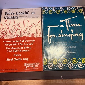 Pode incluir: Dois livros de partituras vintage. O primeiro, "You're Lookin' at Country", apresenta uma capa vermelha com texto branco e uma ilustração de grama. O segundo, "A Time for Singing", tem uma capa azul com escrita branca e redemoinhos decorativos. Ambos são para música vocal.