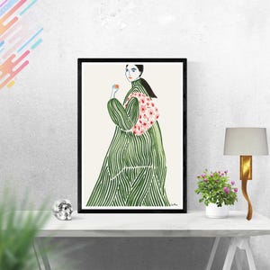 May include: A framed art print showing a watercolour illustration of a person in a long green dress with white stripes and a pink floral shawl. The artwork is displayed on a white table with a lamp and a small potted plant.