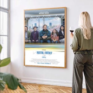 Puede incluir: Un póster enmarcado de la película "Rental Family" con un marco de madera cuelga de una pared blanca. El póster presenta a un grupo de personas en un tren. Una mujer con cabello rubio está cerca, sosteniendo una copa de vino tinto.