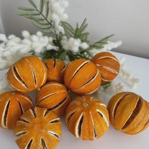 May include: A selection of dried orange slices, displayed on a white surface. The oranges are a warm, golden-orange hue, with visible segments. White and green decorative sprigs are in the background. Suitable for home decor or crafts.