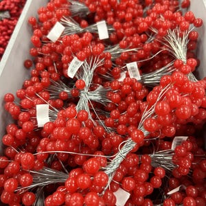 May include: A close-up of bunches of vibrant red berry stems. Each stem has numerous small, round berries, bound with silver wire. The berries are glossy and seem artificial, possibly for decoration. The berries are approximately 1 cm in diameter.