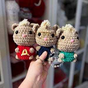 May include: Three handmade crochet chipmunk dolls. Each doll has a tan body, a pink nose, and black eyes. One doll wears a red sweater with a yellow "A", another wears a navy blue sweater and glasses, and the third wears a green sweater.
