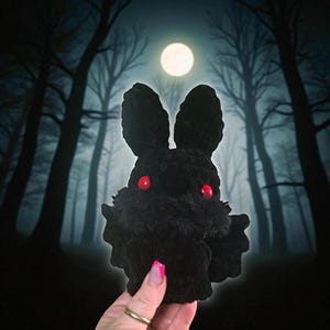 May include: A black crocheted Mothman plushie with red eyes and large ears. The plushie has a fuzzy texture and is set against a backdrop of a moonlit forest. The Mothman is a cryptid from West Virginia.