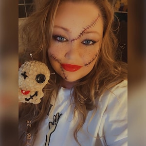 Op de afbeelding: Een persoon met make-up die op steken en sproeten lijkt, houdt een kleine, beige, gehaakte schedel vast met een zwart knoopoog en rode mond. De persoon draagt een wit t-shirt met een zwarte afbeelding en de datum "07 06 '25".