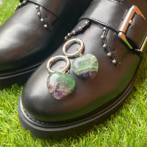 Könnte beinhalten: Schwarze Lederstiefel mit silbernen Schnallen und Nieten. Zwei herzförmige grüne und violette Steine sind mit silbernen Ringen an den Stiefeln befestigt.