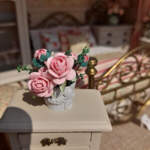 May include: A miniature bedroom scene. A small white nightstand with three drawers and brass handles. A white vase holds pink paper roses with green leaves. The bed has a gold frame and floral bedding.