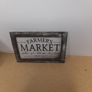 Può includere: Un cartello in legno rustico con sfondo bianco e testo nero che recita "Farmers Market" con le parole "vegetables - fruit - baked goods - dairy - flowers" sotto e "Open Daily" in basso.