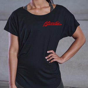 Könnte beinhalten: Schwarzes Kurzarm-Shirt mit dem roten Wort "Blondie" in kursiver Schrift. Das Shirt hat einen weiten Ausschnitt und eine lockere Passform. Darunter ist ein grauer Sport-BH zu sehen.