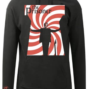 Könnte beinhalten: Schwarzer Kapuzenpullover mit einer Grafik eines Mannes mit ausgestreckten Armen vor einem rot-weißen Spiralhintergrund. Der Text "the Prisoner" steht über dem Bild. Ein kleines rotes Logo befindet sich auf dem linken Ärmel.
