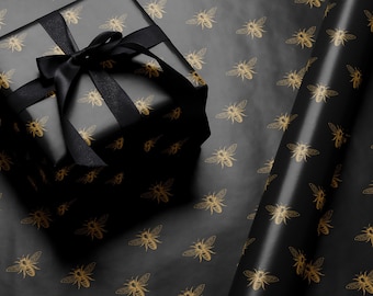 May include: Black gift wrap with a repeating pattern of gold bees. A black gift box with a black ribbon is on top of the wrapping paper.