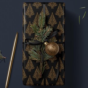 K&ouml;nnte beinhalten: Eine schwarze Geschenkbox, die in goldfarbenes Folienpapier mit einem sich wiederholenden Muster aus Weihnachtsb&auml;umen verpackt ist. Ein Tannenzweig mit einem goldenen Ornament liegt oben auf der Box.