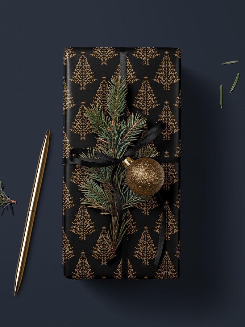 K&ouml;nnte beinhalten: Eine schwarze Geschenkbox, die in goldfarbenes Folienpapier mit einem sich wiederholenden Muster aus Weihnachtsb&auml;umen verpackt ist. Ein Tannenzweig mit einem goldenen Ornament liegt oben auf der Box.