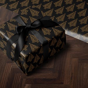 K&ouml;nnte beinhalten: Eine schwarze Geschenkbox mit einem goldenen Weihnachtsbaum-Muster und einem schwarzen Satinband. Die Box steht auf einem dunklen Holzboden.