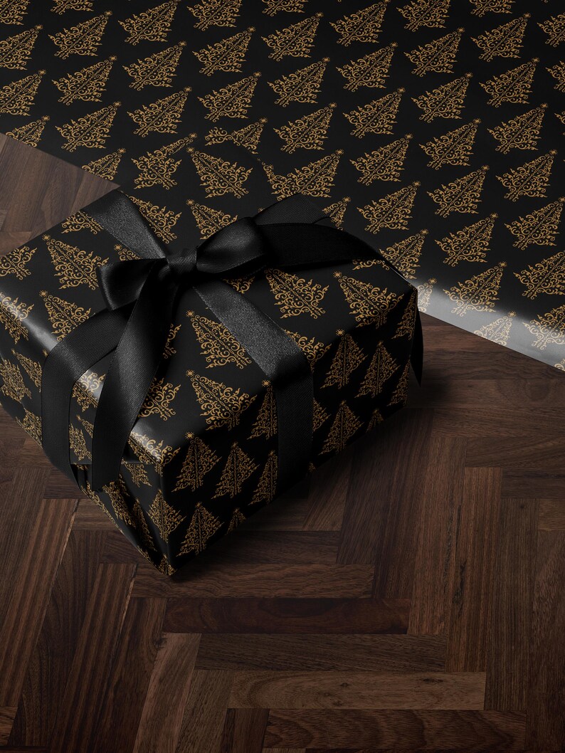 K&ouml;nnte beinhalten: Eine schwarze Geschenkbox mit einem goldenen Weihnachtsbaum-Muster und einem schwarzen Satinband. Die Box steht auf einem dunklen Holzboden.