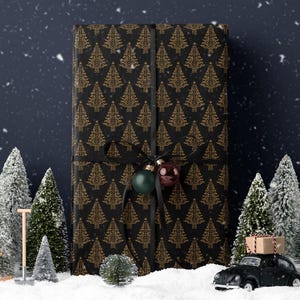 K&ouml;nnte beinhalten: Eine schwarze Geschenkbox mit einem goldenen Weihnachtsbaum-Muster, umwickelt mit einem schwarzen Band und geschm&uuml;ckt mit gr&uuml;nen und roten Kugeln. Die Box steht in einer verschneiten Szenerie mit Miniatur-Kiefern und einem schwarzen Spielzeugauto.