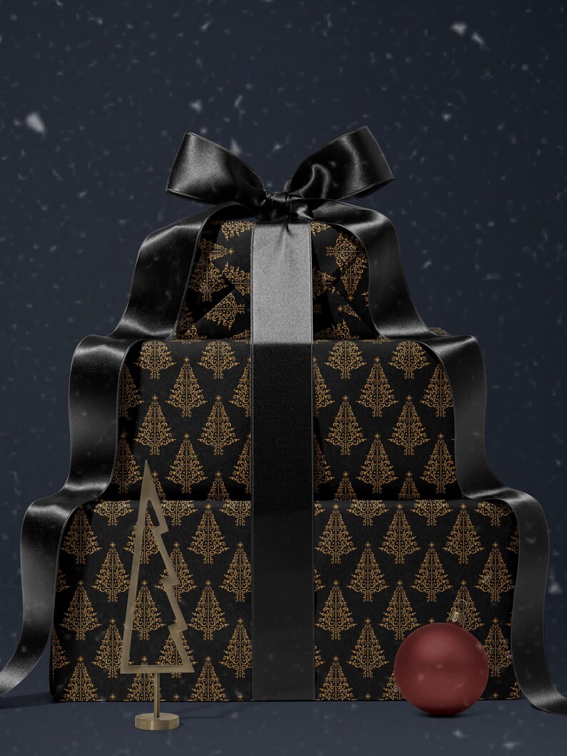 K&ouml;nnte beinhalten: Drei schwarze Geschenkboxen, die &uuml;bereinander gestapelt sind und in schwarzes Papier mit einem goldenen Weihnachtsbaum-Muster eingewickelt sind. Die oberste Box hat ein schwarzes Satinband, das zu einer Schleife gebunden ist. Ein goldener Weihnachtsbaum-Anh&auml;nger aus Metall befindet sich vor den Boxen, und ein roter Anh&auml;nger befindet sich auf der untersten Box.