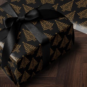 K&ouml;nnte beinhalten: Eine schwarze Geschenkbox mit einem schwarzen Band und einem goldenen Weihnachtsbaum-Muster. Die Box steht auf einer dunklen Holzoberfl&auml;che.