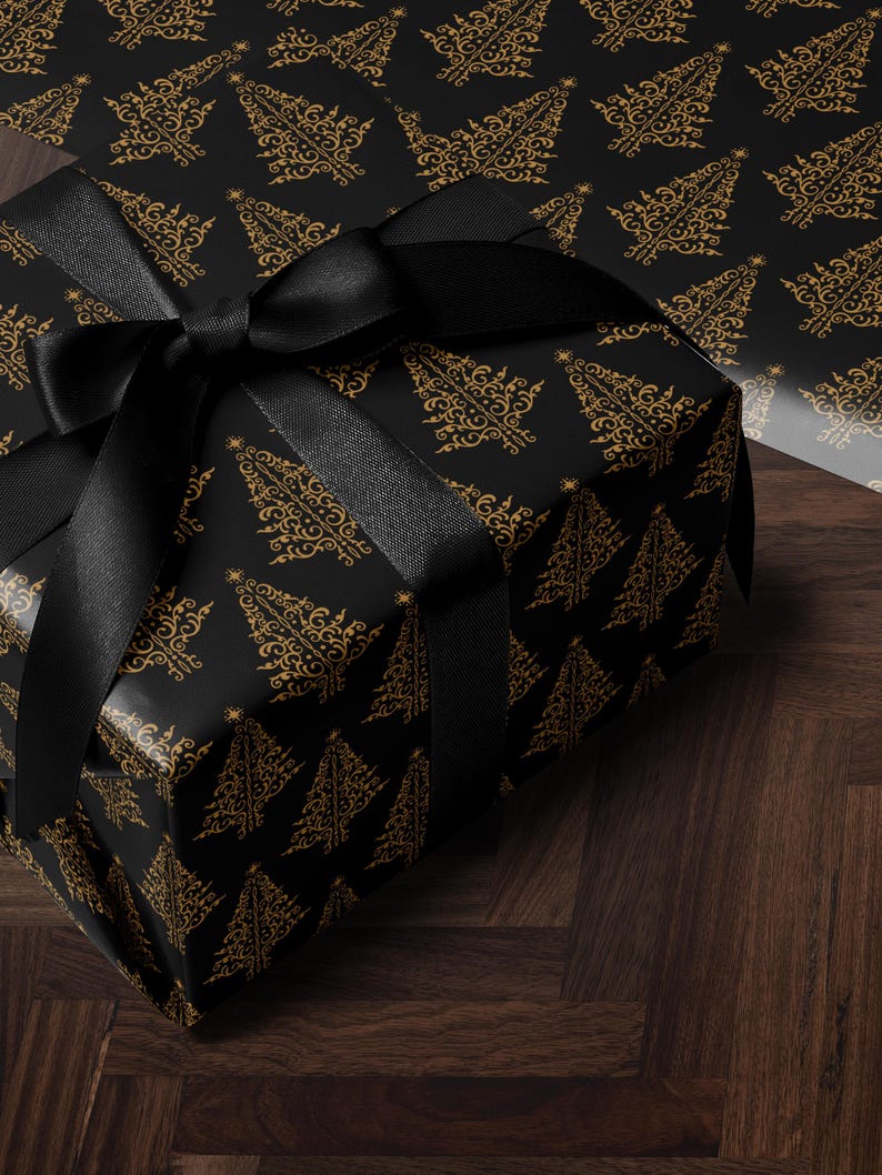 K&ouml;nnte beinhalten: Eine schwarze Geschenkbox mit einem schwarzen Band und einem goldenen Weihnachtsbaum-Muster. Die Box steht auf einer dunklen Holzoberfl&auml;che.