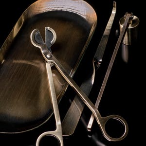May include: A set of brass-toned metal tools including a pair of wick trimmers with circular handles, a long slender wick dipper with a flat end, and a small cylindrical candle snuffer, all arranged alongside an oval-shaped metal tray.