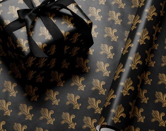 Puede incluir: Papel de regalo negro con un patrón de flor de lis dorado. Una caja de regalo negra con una cinta negra está envuelta en el papel. También se muestra un rollo de papel de regalo.