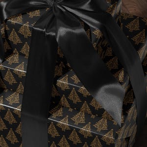 K&ouml;nnte beinhalten: Ein Stapel von drei schwarzen Geschenkboxen mit goldenen Weihnachtsbaummustern. Die Boxen sind mit einem schwarzen Satinband gebunden.