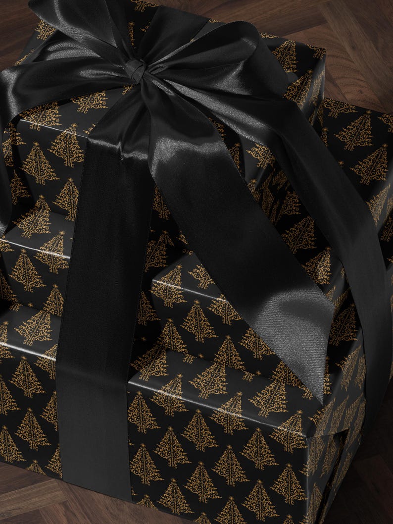 K&ouml;nnte beinhalten: Ein Stapel von drei schwarzen Geschenkboxen mit goldenen Weihnachtsbaummustern. Die Boxen sind mit einem schwarzen Satinband gebunden.