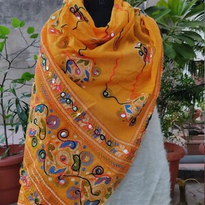 K&ouml;nnte beinhalten: Ein gelber Seidenschal mit aufwendigen Stickereien und Spiegelarbeiten. Der Schal zeigt ein geometrisches Muster mit floralen Motiven in Blaut&ouml;nen, Gr&uuml;n, Rot und Orange.