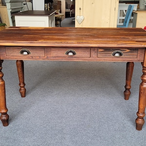 Könnte beinhalten: Ein dunkelbrauner Holztisch mit drei Schubladen und Metallgriffen. Der Tisch hat vier gedrehte Beine. Die Abmessungen des Tisches eignen sich für ein Ess- oder Wohnzimmer. Der Tisch ist etwa 150 cm lang.