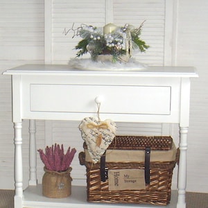 Könnte beinhalten: Weißer Konsolentisch mit einer Schublade und einem unteren Regal. Der Tisch hat gedrehte Beine und eine weiße Lackierung. Ein Weidenkorb mit einem Stoffdeckel und dem Text "My Home Storage" steht auf dem Regal. Eine kleine Vase mit Trockenblumen und eine herzförmige Dekoration stehen ebenfalls auf dem Regal.