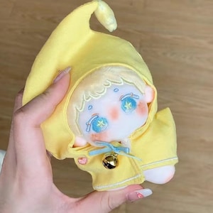 May include: A yellow plush toy with a pointed hood, embroidered eyes, and a bell. The toy is wearing a yellow jacket with white trim.