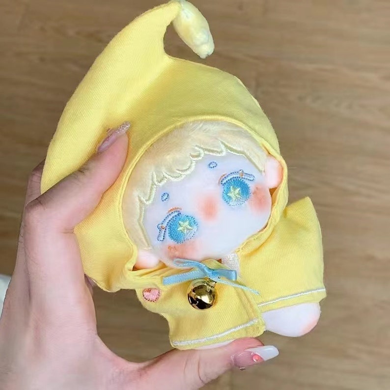 May include: A yellow plush toy with a pointed hood, embroidered eyes, and a bell. The toy is wearing a yellow jacket with white trim.