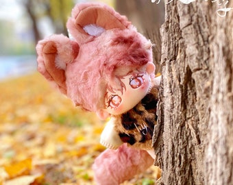 Könnte beinhalten: Ein Plüschtier mit einem rosa Fellhut und einer Jacke mit Leopardenmuster. Das Spielzeug schaut hinter einem Baumstamm hervor. Der Hintergrund ist ein verschwommenes Bild von Herbstblättern.