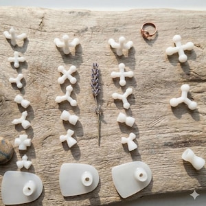 May include: A collection of white massage tools and a copper ring are displayed on a weathered wooden surface. The tools are in various shapes, including crosses, Ys, and rounded rectangles. A sprig of lavender is also present.