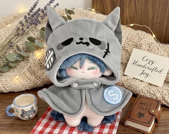 May include: A plush doll wearing a gray hooded cape with cat ears, blue hair, and rosy cheeks. The doll is on a red and white checkered cloth, with a small teacup and a miniature book that says "WOLF TALES". A card reads "Cozy Handcrafted Joy".