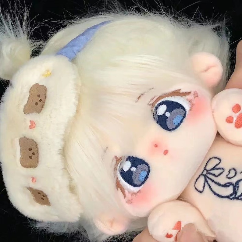 May include: A plush doll with blonde hair, blue eyes, and a white furry eye mask with brown bear appliques. The doll is wearing a white shirt with a floral design and has a pink blush on its cheeks.