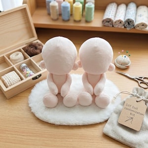 May include: Two plush, light pink doll bodies with rounded heads and small limbs sit on a white, fluffy mat. A wooden box with craft supplies, spools of thread, and a DIY doll customization kit are also visible, suggesting a craft project.