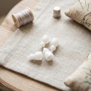 Op de afbeelding: Een verzameling witte plastic naaibenodigdheden, waaronder vier kleine, kalebasvormige stukken, een vingerhoed en een spoel garen met roze en blauw garen. De items zijn gerangschikt op een lichtgrijze linnen ondergrond, met een speldenkussen op de achtergrond.