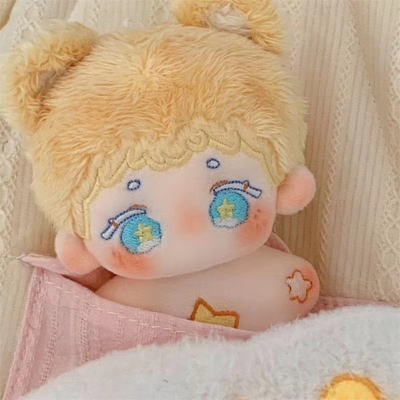 May include: A plush toy with blonde hair, blue eyes, and a pink and white blanket. The toy has a star design on its chest and a flower design on its arm.