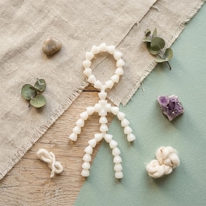 May include: A white, articulated figure in a human shape, composed of connected heart-shaped pieces, lies on a wooden surface. The figure is surrounded by natural elements, including a stone, eucalyptus leaves, a purple crystal, and a ball of white fiber.