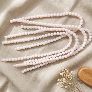 May include: Several strands of light pink square beads are arranged on a beige fabric. The beads are uniform in size and shape, creating a delicate and elegant appearance. A small wooden object and dried flowers are in the lower right corner.
