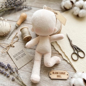 Peut inclure: Une poupée en tissu rose clair, vierge, avec une tête ronde et de petits bras et jambes. Des fournitures de couture, dont une bobine de fil, des ciseaux et un coussin à épingles, entourent la poupée. Une étiquette indique "HANDMADE WITH LOVE" et une autre "BLANK CANVA' DOLL".