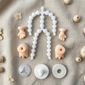 May include: A collection of doll-making supplies, including a white articulated skeleton, small hands, feet, and various round components. The pieces are arranged on a neutral-toned fabric background, suggesting a craft or hobby theme.