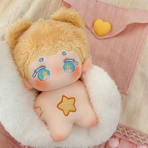 May include: A plush toy with blonde hair, blue eyes, and a star on its chest. The toy is wearing a white fluffy coat and is sitting on a pink and white background.