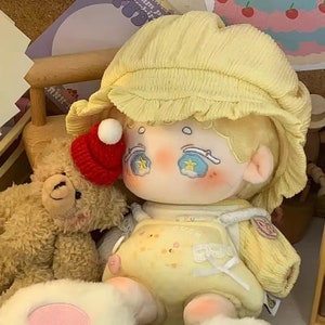 May include: A plush toy doll with a yellow hat and a yellow and white outfit. The doll has a red hat on its head and is holding a small teddy bear.