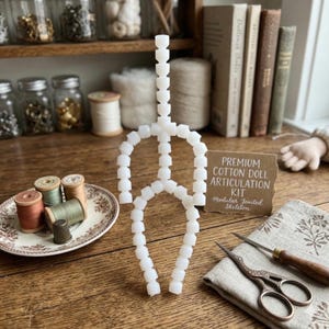 Könnte beinhalten: Ein weißes, modulares Puppenskelett steht auf einer Holzoberfläche, neben einem Teller mit Garnrollen und einem Fingerhut. Eine Karte trägt die Aufschrift "Premium Cotton Doll Articulation Kit". Schere und ein Nähwerkzeug sind ebenfalls vorhanden.
