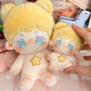 May include: Two plush toys shaped like children with yellow hair, blue eyes, and a star design on their chests. The larger toy is standing and the smaller toy is sitting.