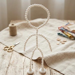 May include: A white articulated figure, resembling a stick figure, made of heart-shaped beads. The figure stands on a wooden surface, with a spool of thread and scissors in the background. The figure's arms and legs are curved.