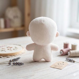 May include: A plush doll body in a light pink color, with a rounded head, small ears, and stubby arms and legs. The doll is positioned on a light-colored wooden surface, with spools of thread and lavender sprigs nearby. A tag reads "Handmade with Love."