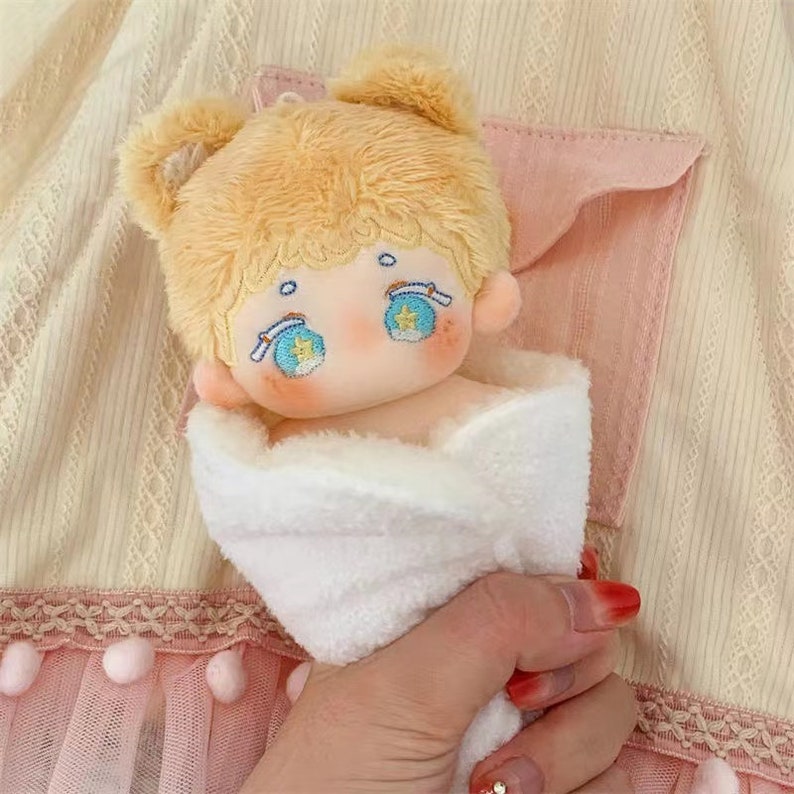 May include: A plush toy doll with blonde hair, blue eyes, and rosy cheeks is wrapped in a white blanket. The doll is wearing a white headband with blue and yellow stars.