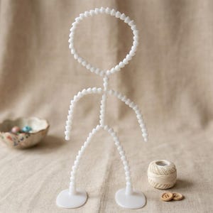 May include: A white articulated figure with a circular head and jointed limbs, standing on two circular bases. The figure is made of small, connected pieces. A small bowl with colorful beads, a spool of thread, and two wooden buttons are also present.